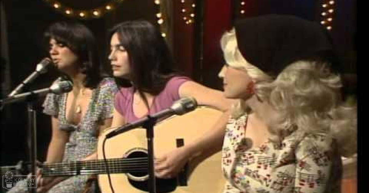 A young adult female singer in a patterned dress performs alongside other musicians in an indoor setting with string lights and warm lighting, suggesting an evening live music venue.
