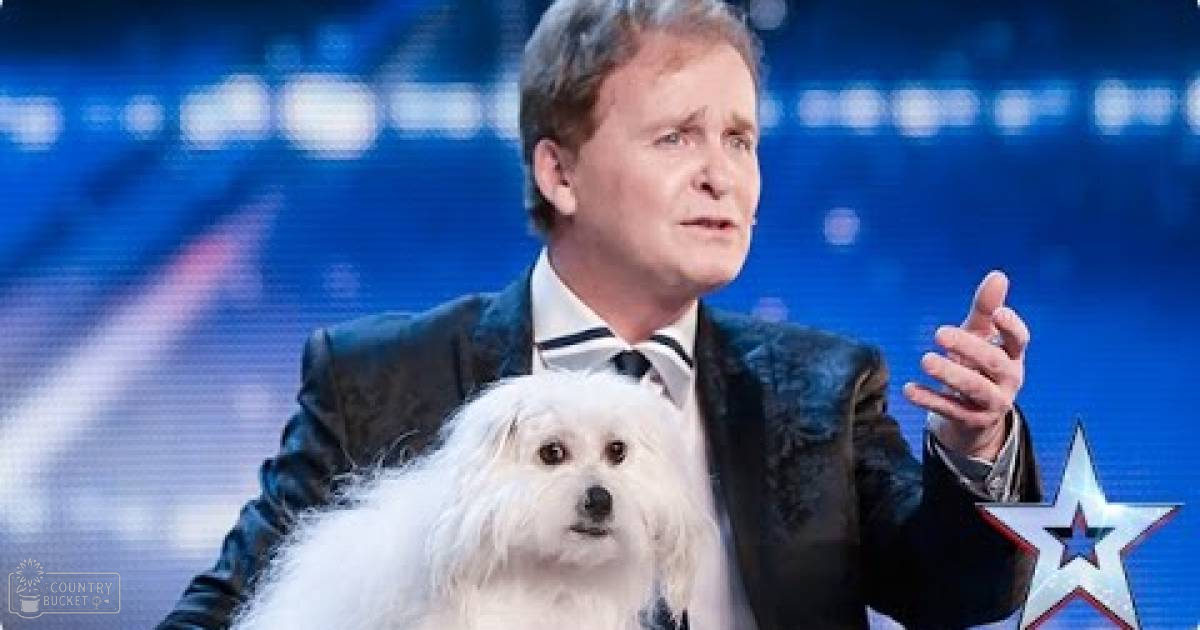 A Maltese dog with white fur and an adult male performer in a dark blazer over a white shirt and black tie, likely at a talent show under bright blue stage lighting.