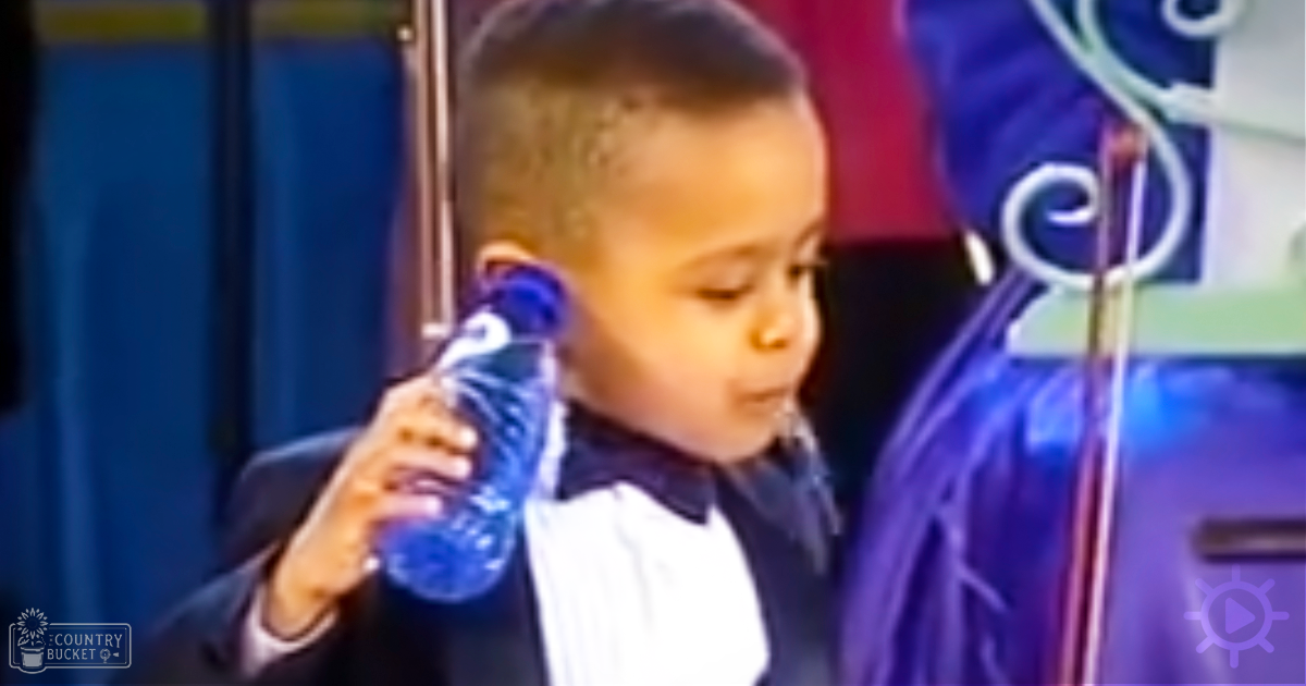 A young boy in a dark suit with a white shirt and black bow tie stands indoors, holding a blue water bottle near a microphone stand.