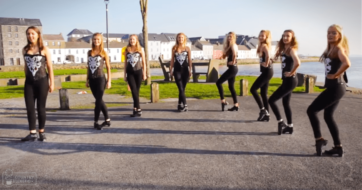 Young adult females performing a dance routine on an outdoor waterfront promenade at dusk, surrounded by greenery and residential structures.