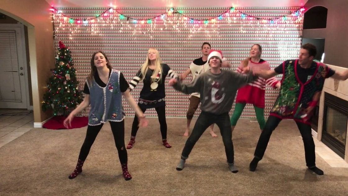 Young adults dancing in a festive Christmas living room, wearing holiday-themed outfits with red boots and patterned vests.