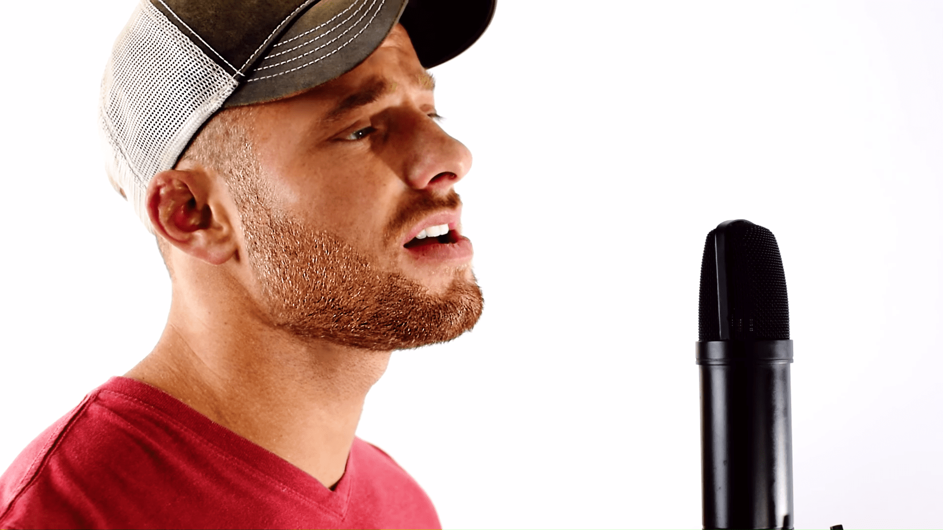 A young adult male wearing a bright red, relaxed fit t-shirt stands indoors against a plain white background. A black microphone with a foam cover is slightly out of focus to the right.