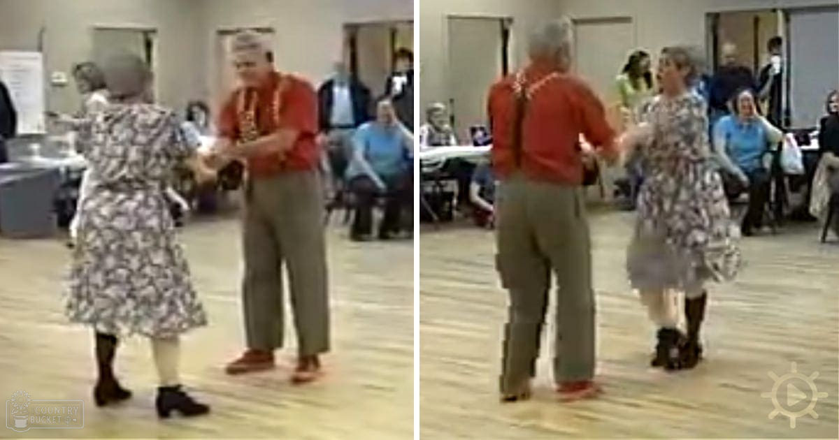 An older couple dancing in a community center, surrounded by tables with chairs, under natural light during the daytime.