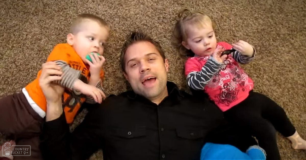 Middle-aged adult male wearing a black button-up shirt indoors with two toddlers in colorful outfits nearby on a carpeted floor during the daytime.