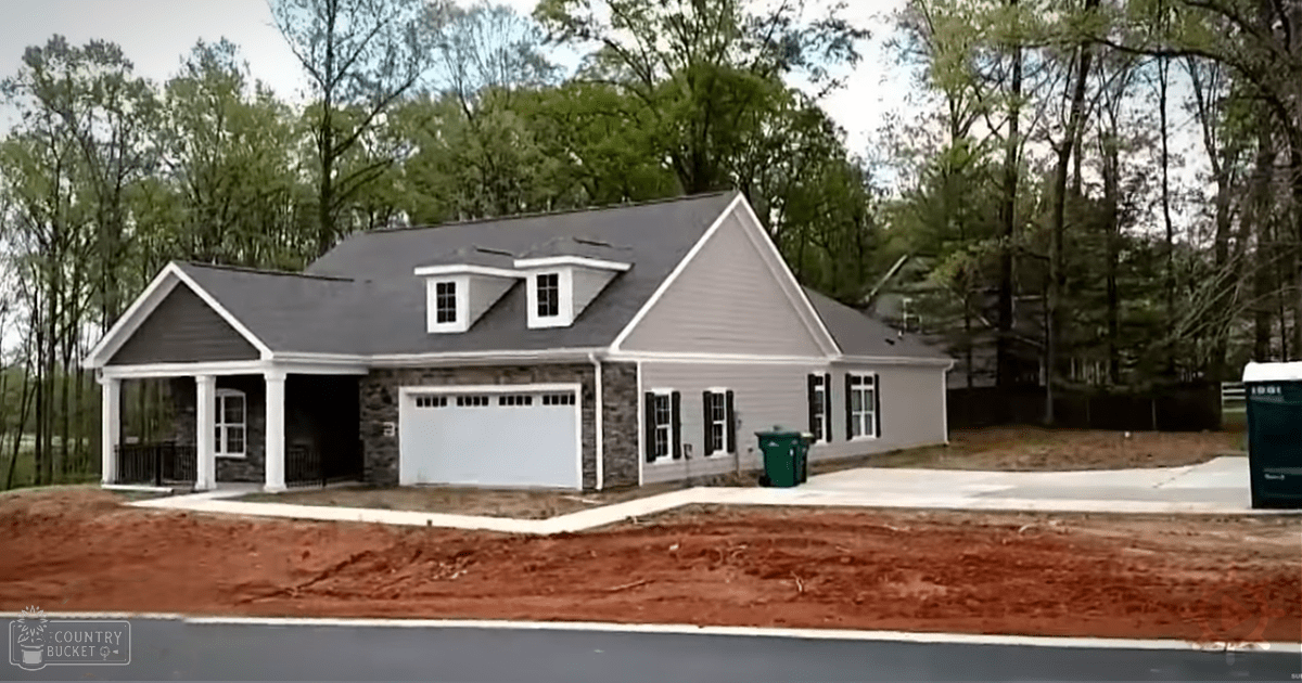 A newly built suburban house with gray siding, white trim, a garage door, a small porch area with columns, and a driveway; bare soil and leafless tree branches suggest it's not spring or summer.