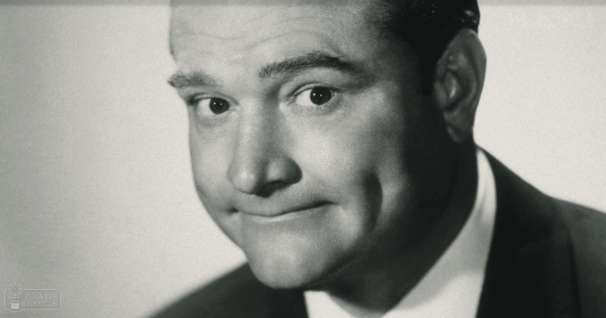 An adult male in a professional studio setting, wearing a dark-colored suit jacket over a white dress shirt.