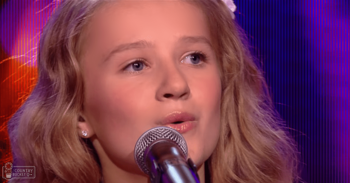 A young female child wearing earrings sings into a microphone against a stage backdrop with vibrant purple and blue lighting.