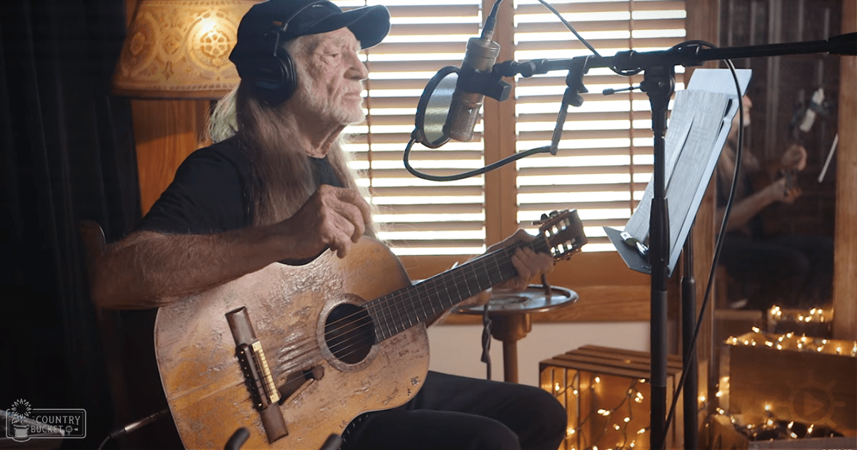 An older adult male musician in a home recording studio, wearing a black t-shirt and dark pants with headphones around the neck, surrounded by musical equipment under natural light filtering through wooden blinds.