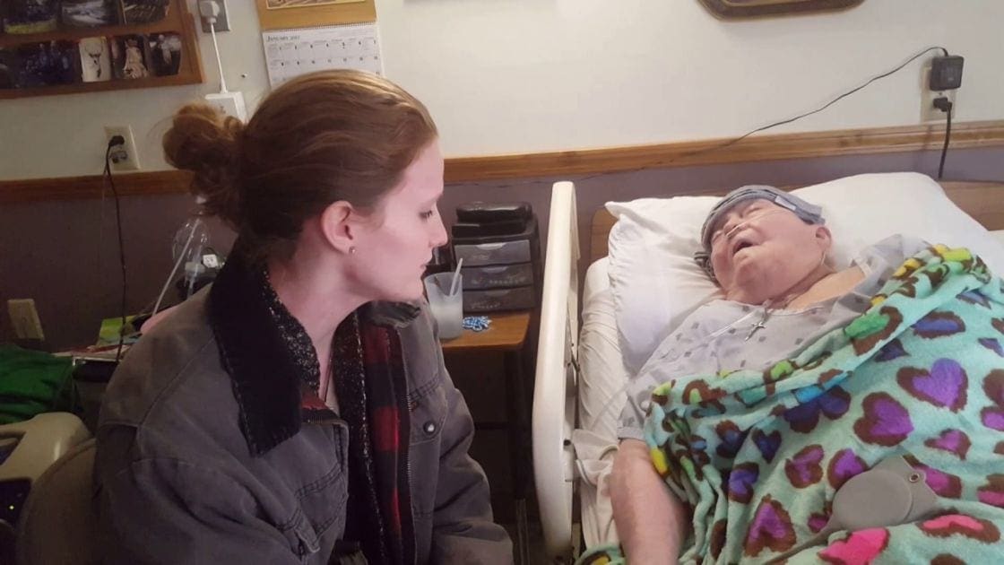 A young adult female and an older adult in a hospital room with medical furniture and personal items on the bedside table.