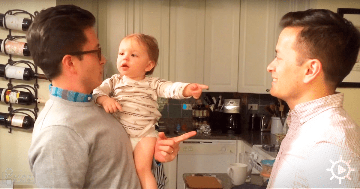 A baby in white onesie points to right, held by adults in kitchen setting at dusk.