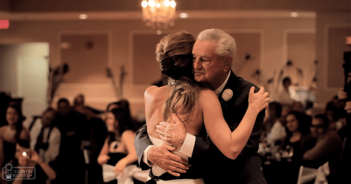 An older couple dancing closely at an indoor wedding reception, surrounded by guests seated and clapping under chandeliers with warm ambient lighting.