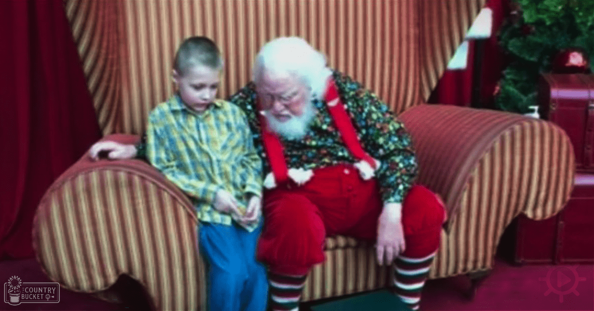 An older adult male Santa Claus with a white beard and colorful patterned shirt stands indoors among festive decorations, including red curtains and potted plants, with a child nearby wearing a yellow-green plaid shirt in the foreground.