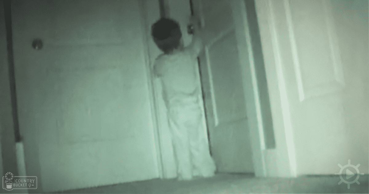 A toddler-sized child wearing light-colored pants and a white shirt stands indoors near a door frame.