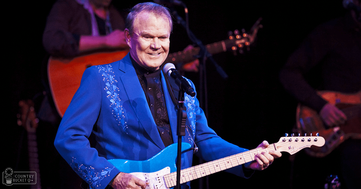 A middle-aged male performer wearing a blue suit jacket with floral embroidery and black shirt, standing on an indoor concert stage surrounded by other musicians.