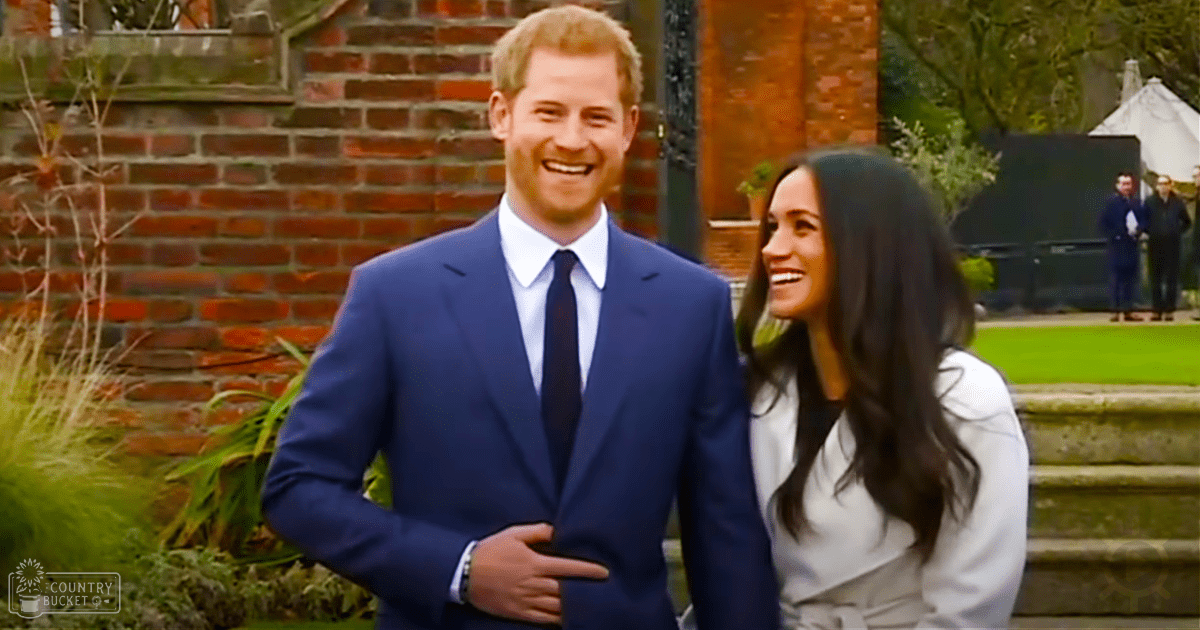 A couple in formal attire stands outdoors near a white tent structure with brick walls and greenery in the background.