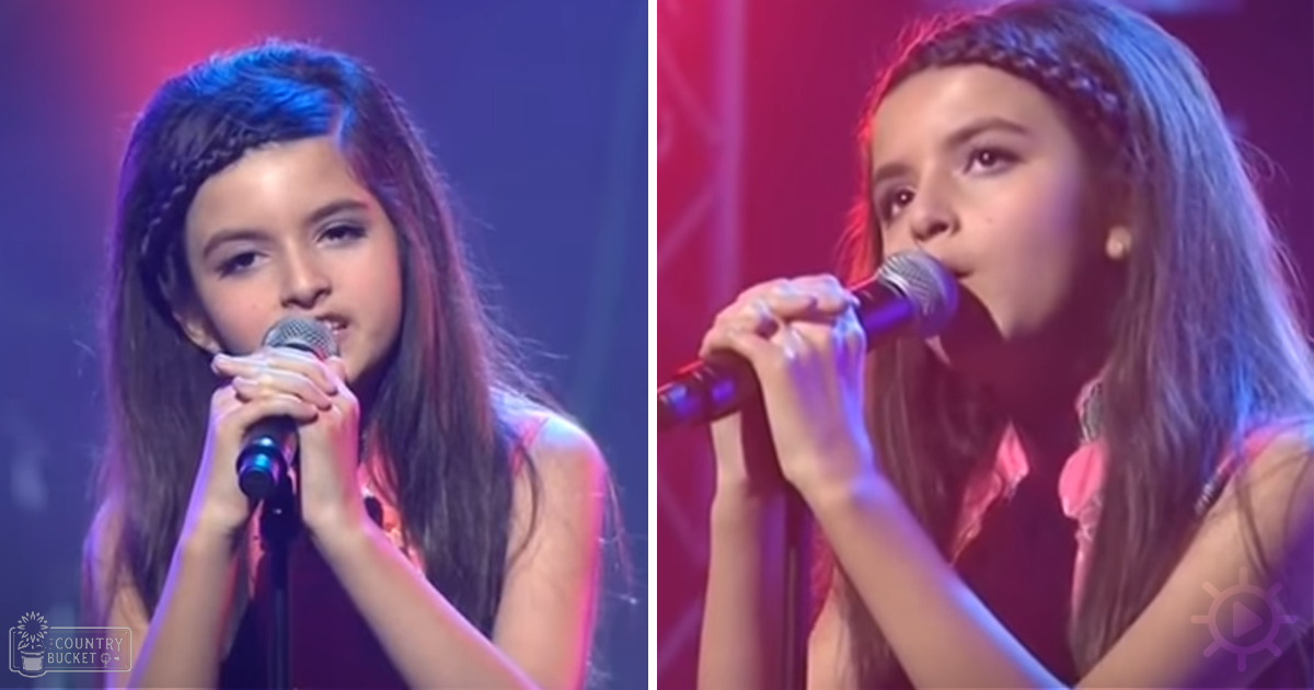 A young female child performs on an indoor stage with red and blue lighting, wearing a sleeveless pinkish-orange top with subtle floral patterns.