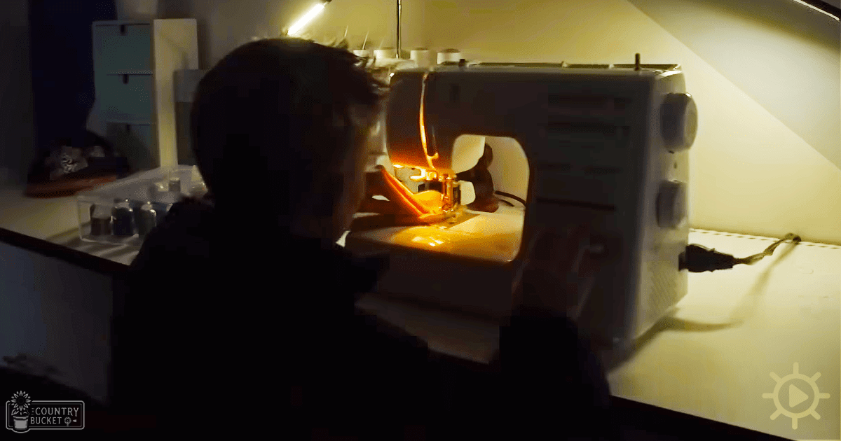A sewing machine in use, illuminated by a light above it, surrounded by various small items, possibly in an indoor home or workshop setting with beige walls and yellowish lighting.