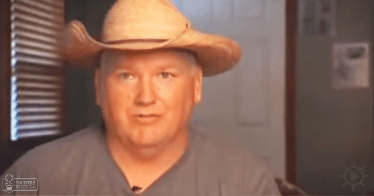 An older adult male wearing a light brown cowboy hat with a wide brim, standing indoors near a partially visible window with blinds and wall decorations in neutral tones.