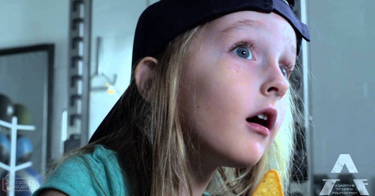 A young adult female with long blonde hair, wearing a black baseball cap backward and a teal t-shirt indoors.