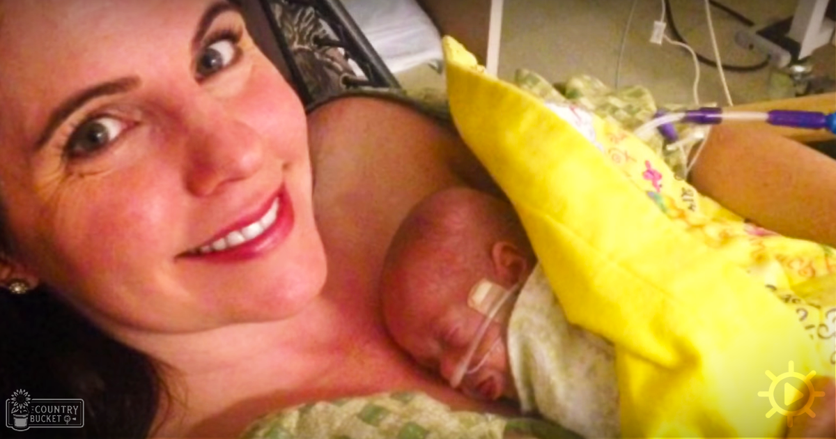 A woman holding an infant in a hospital setting, wearing a dark-patterned garment and smiling; medical tubes visible attached to baby's head.