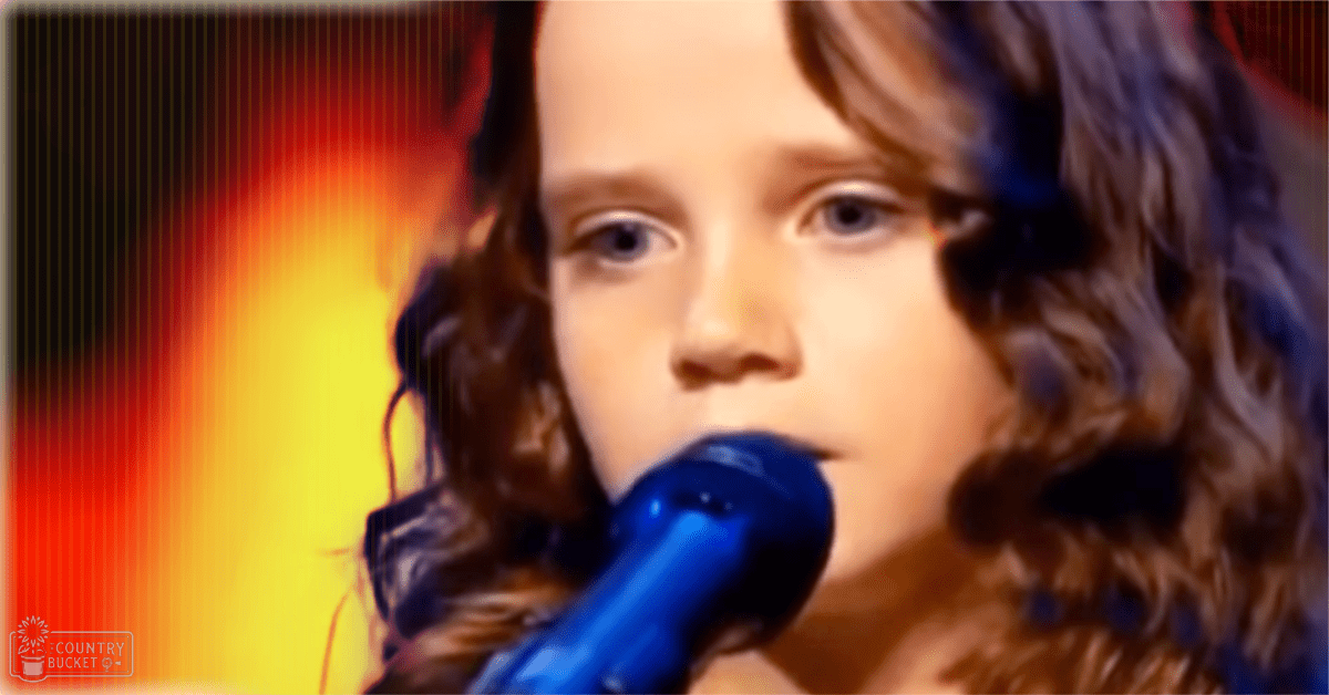 A toddler/child wearing a dark textured garment sings into a blue microphone on stage under warm lighting, suggesting an evening talent show performance.