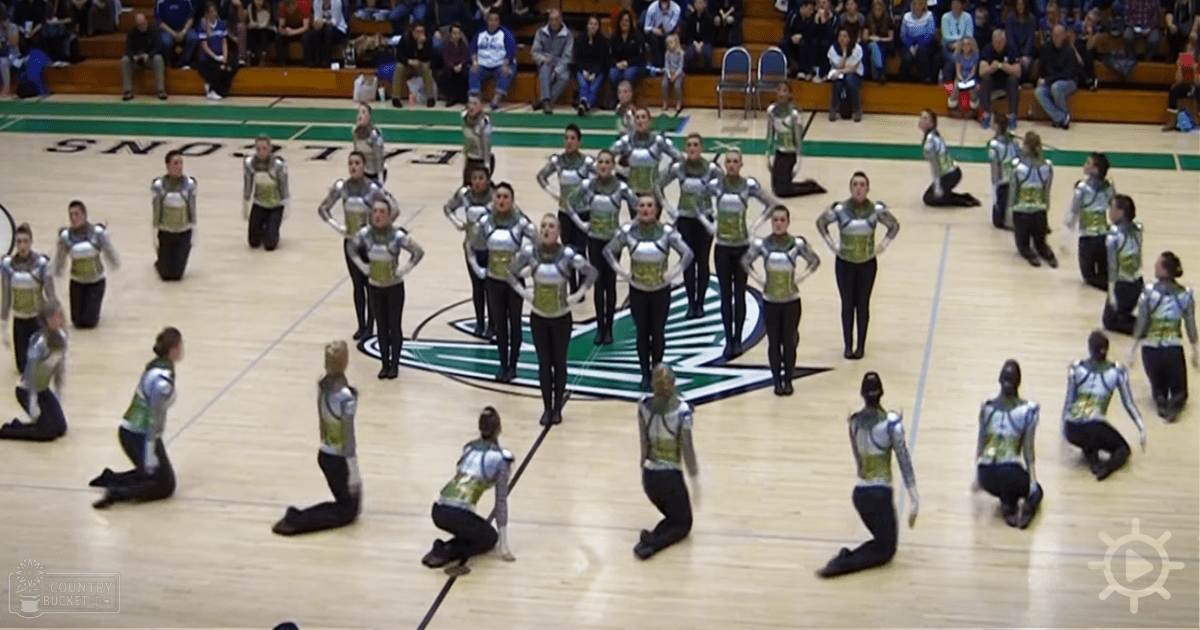 A dance team of young adult females performing in form-fitting, silver and green metallic armor-like leotards at an indoor sports arena with spectators seated on bleachers.