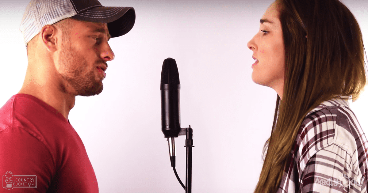 A duet recording session in an indoor studio setting with two young adults, one wearing a red t-shirt and the other in a plaid shirt. A black microphone is prominently featured between them against a plain white background.