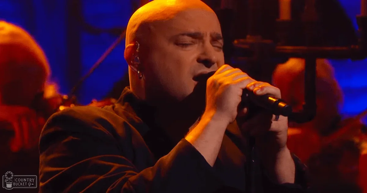 An adult male singer wearing a dark long-sleeved V-neck garment at a concert with stage lights in the background.