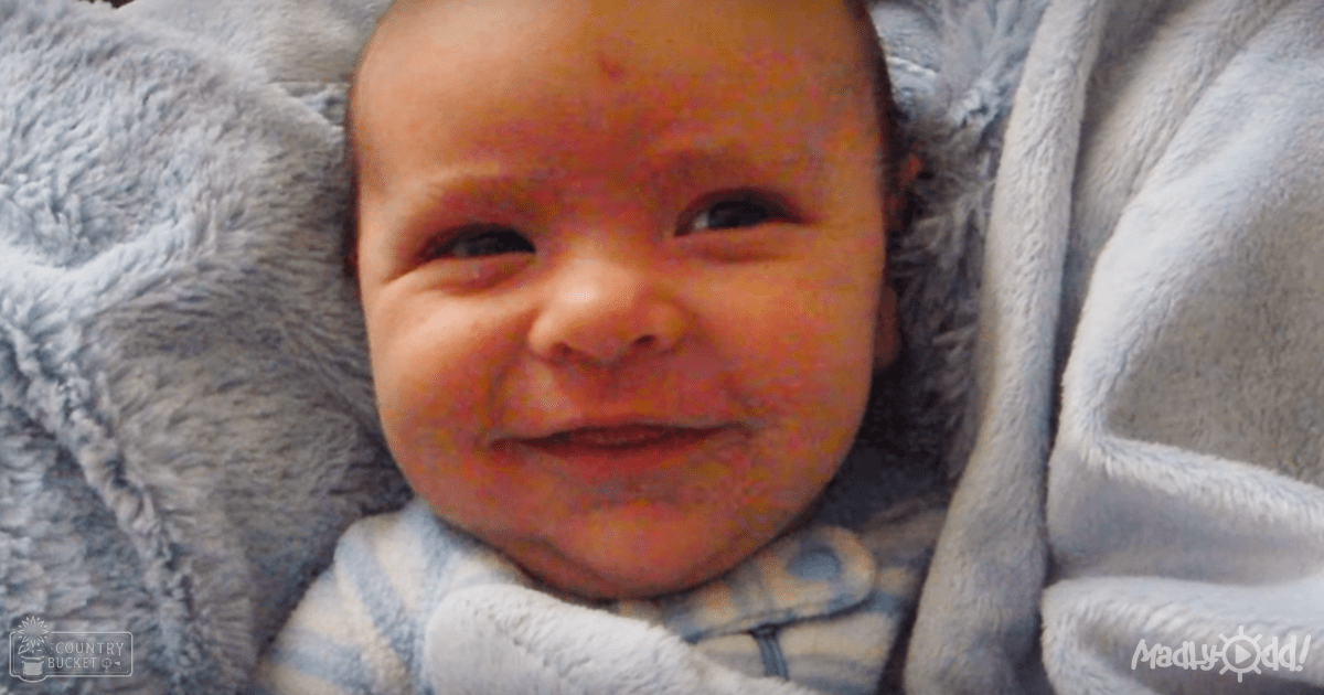 A baby wearing a light blue and white striped onesie with fur-like trim, lying indoors under a gray plush blanket.