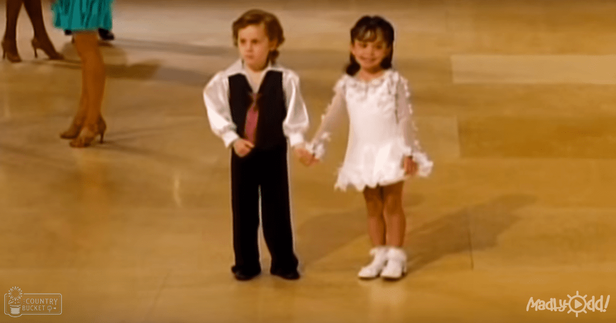 Indoor dance floor setting with two children holding hands, wearing vintage-inspired white outfits and pink tie; light reflecting off polished wooden floor suggests evening or night lighting.