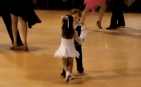 A toddler in a white ruffled dress dances with an adult in a dance studio, surrounded by other dancers and wooden floors under artificial lighting.