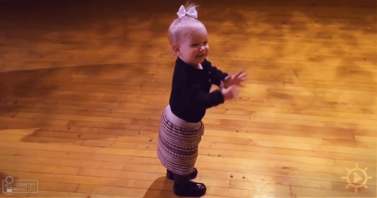 A toddler wearing a dark long-sleeve top, gray and white striped skirt, black shoes, and a white bow on her head, standing indoors with polished wooden flooring.