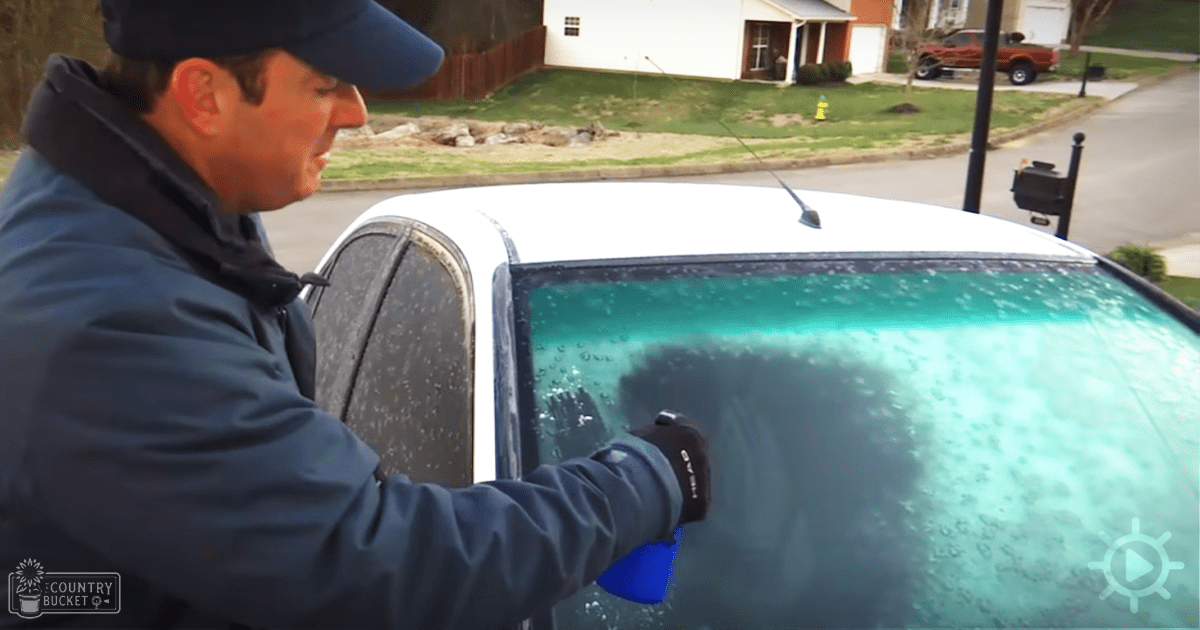 An adult male wearing a dark blue, relaxed-fit, collarless jacket cleaning his white car in a residential neighborhood with greenery visible on the grassy area.