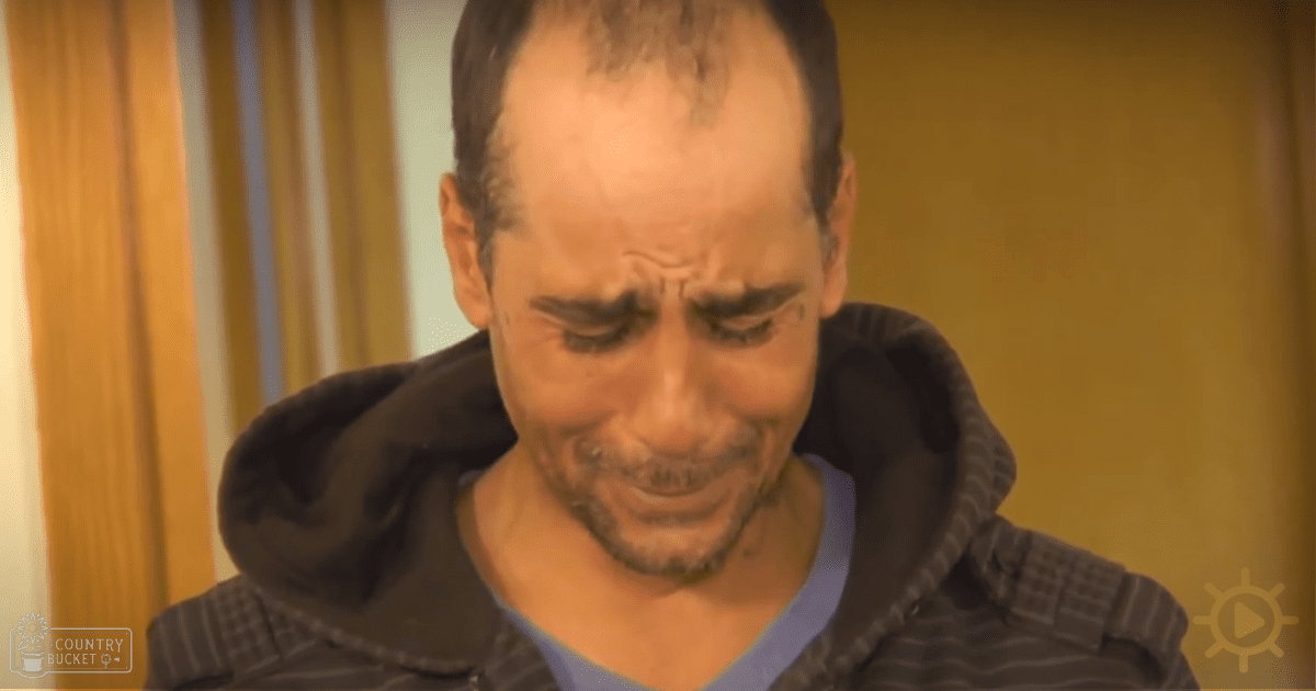 Middle-aged man wearing a dark hoodie with striped sleeves and a light blue v-neck shirt, standing indoors against wooden paneling or curtains.