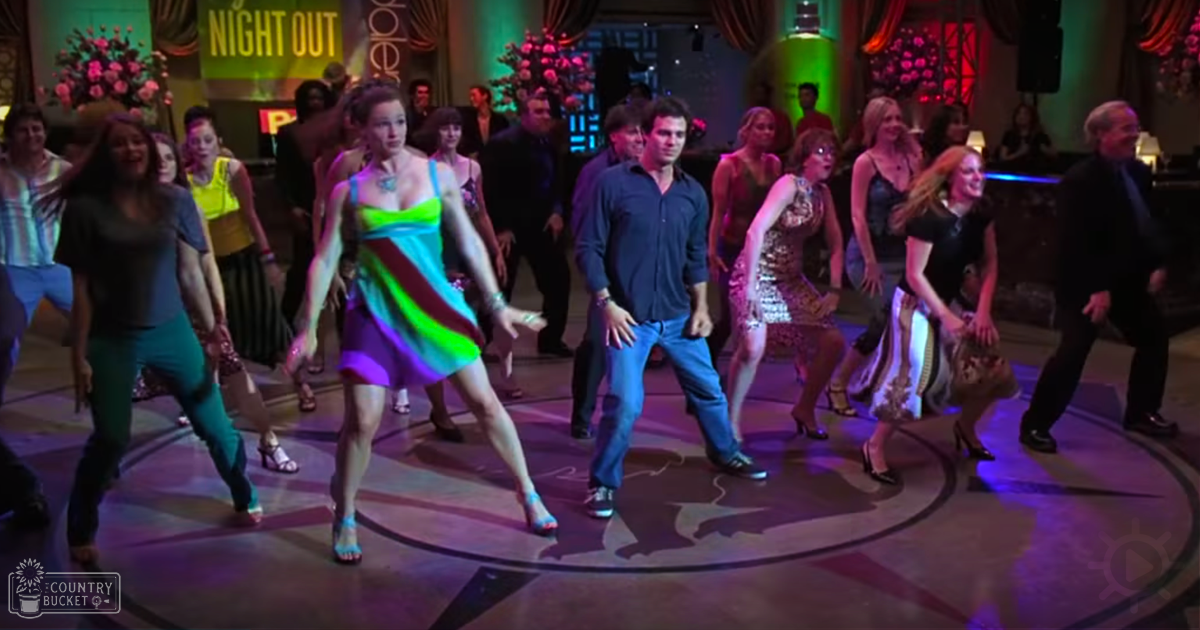 A lively dance scene at an indoor nightclub or party venue, with young adults in vibrant outfits against decorative floral arrangements and dimly lit stage lights.