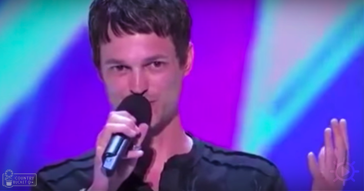 A young adult male in a dark, textured shirt performing on stage under vibrant lighting effects.