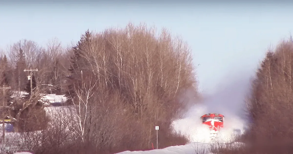 A snowy rural landscape with a red train emitting smoke, leafless trees, and a clear sky in winter; snow-covered ground and a signpost with an arrow on the left.