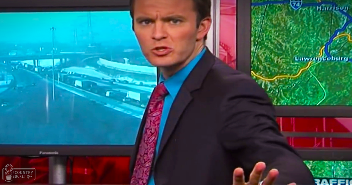A news anchor wearing a dark suit jacket and light blue shirt with a patterned tie in a professional studio setting displaying two monitors showing traffic and geographical information.