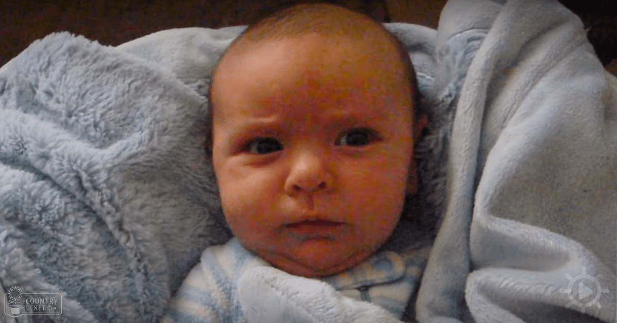 A baby wrapped in a light blue blanket, lying on a textured grayish-blue surface indoors with soft natural lighting.