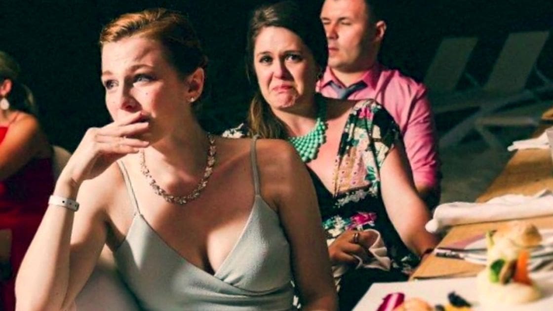 A young adult woman in a light gray dress with thin straps stands at a table set for dinner, surrounded by other guests dressed formally; indoor setting suggests evening event.