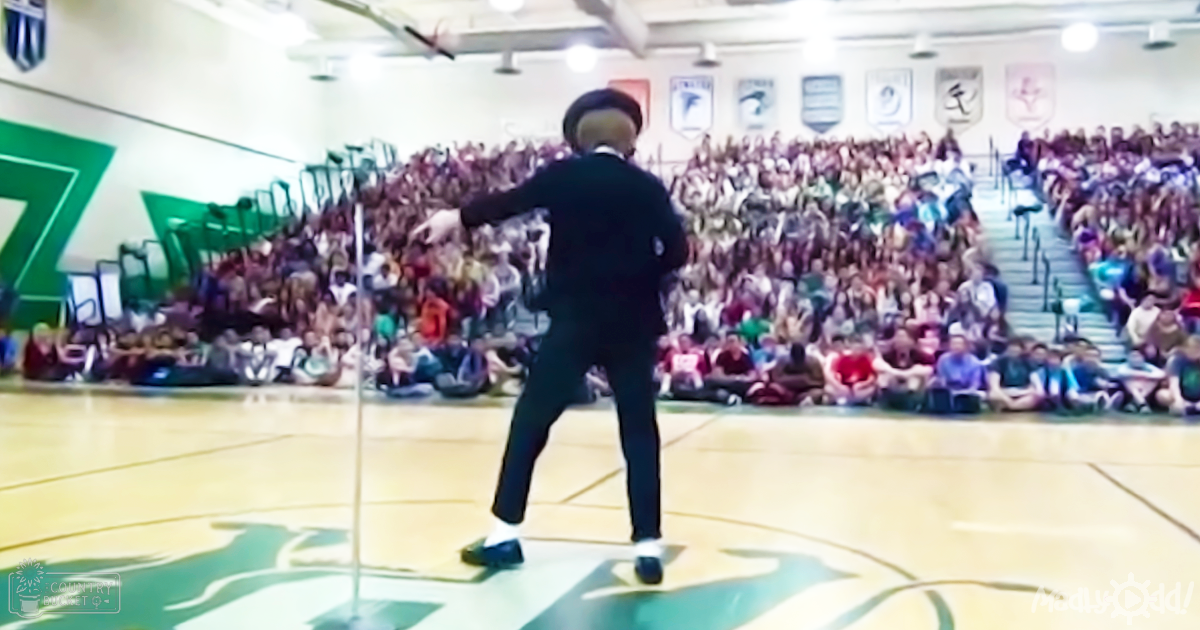 An adult male in a dark suit dancing at a school gymnasium with children and young adults seated on bleachers, dressed casually.