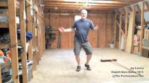A middle-aged adult male in a relaxed fit, dark gray t-shirt with a logo on the left chest area and dark green shorts stands amidst an unfinished basement setting with exposed wooden beams and insulation. Various tools and containers are scattered around.