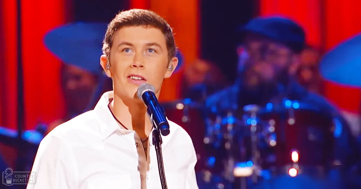 A young adult male singer wearing a white dress shirt with an earpiece microphone, performing on stage at 'The Voice' event. The background features musical equipment and red stage lighting.
