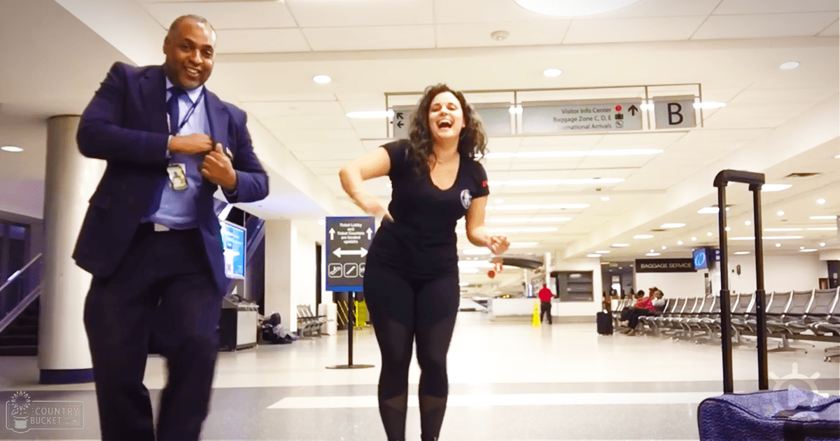 Adult female in an airport setting, wearing a black t-shirt with a logo and dark leggings. Adult male in a dark blue suit jacket smiles beside her.