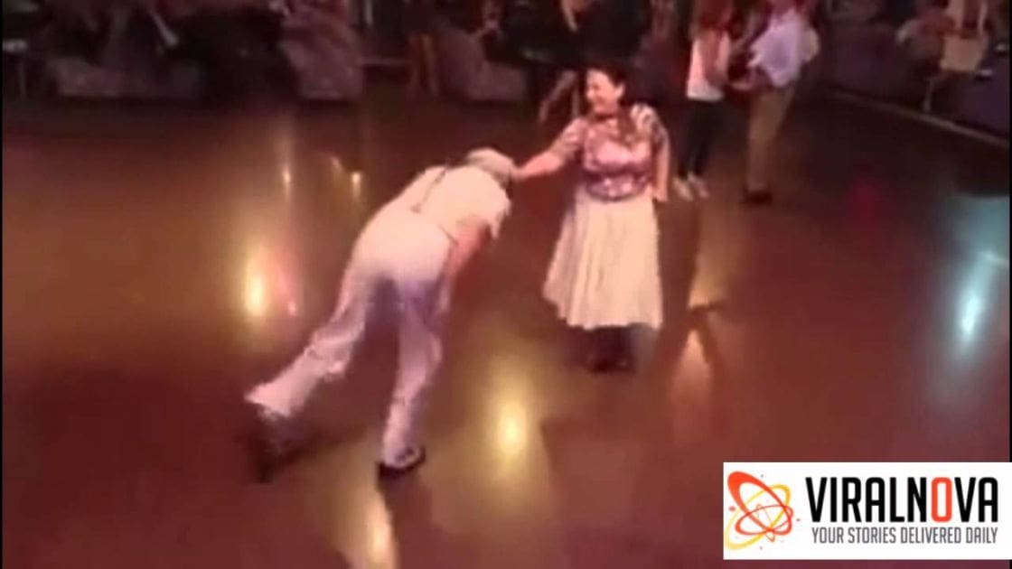 An adult male dancer wearing light-colored ruffled trousers and a white shirt, paired with an adult female dancer in a pink patterned blouse and white skirt, on a polished wooden dance floor indoors under artificial evening lighting.