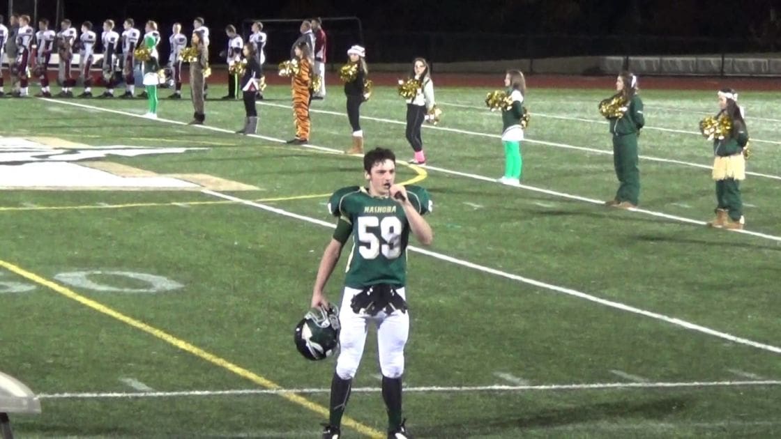 A young adult male football player stands on a high school football field at night during fall, surrounded by cheerleaders performing routines.