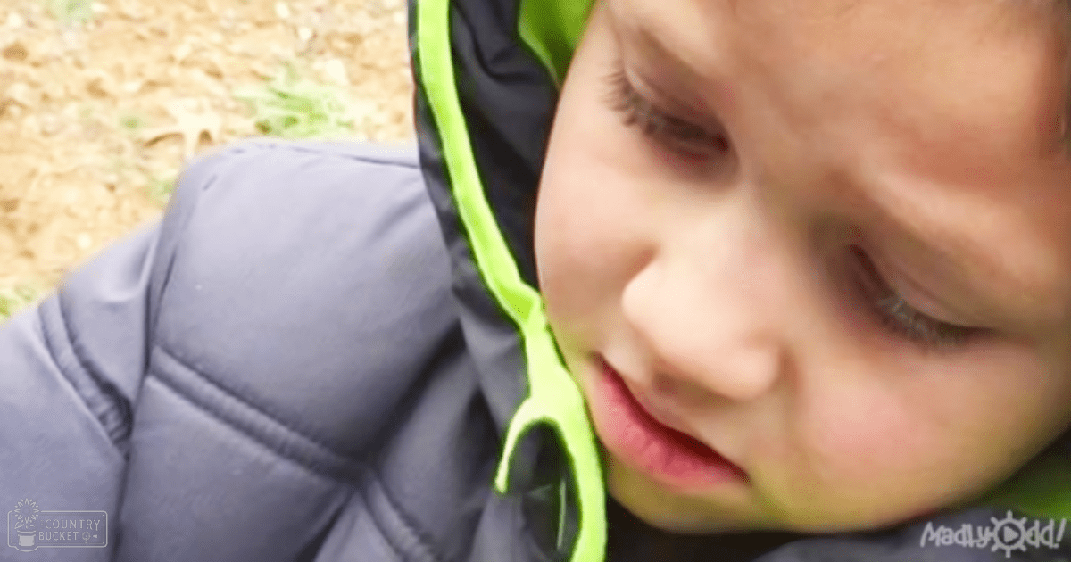 A toddler in a park setting wearing a dark blue puffer jacket with neon green accents, surrounded by greenery under bright daylight.
