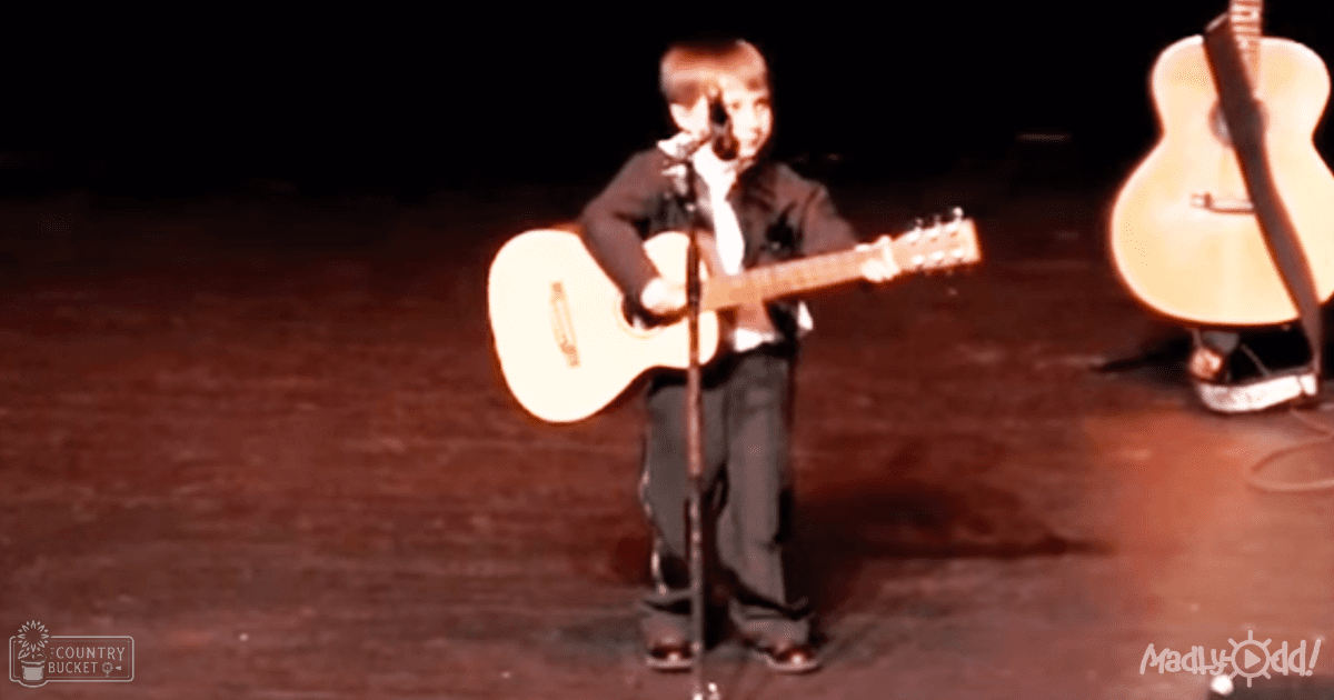 A young adult male in a dark suit jacket and white shirt plays guitar on stage, with a microphone stand and another guitar visible in the background.