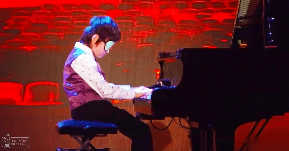 A young adult male in a white shirt with black polka dots and glasses plays the piano in an indoor concert setting, surrounded by red seats under stage lights.