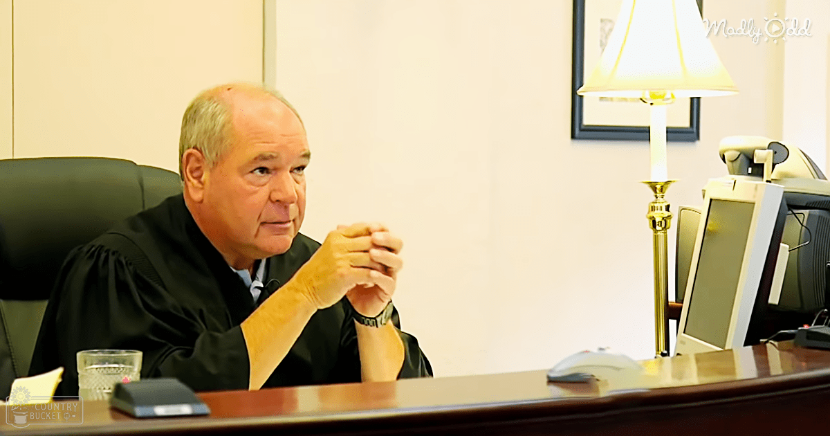 A middle-aged man in a black judicial robe seated at a desk with various items including a computer monitor, lamp, glass of water, notepad, phone, and microphone or intercom device in an indoor courtroom setting.